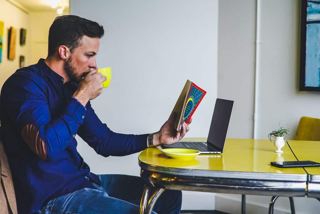 profile view of man sipping coffee from yellow mug whilst reading book sat at yellow desk with laptop