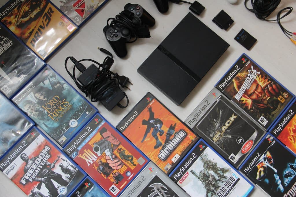 offset top down view of playstation 2 video games on a white table alongside a ps2 slim console, controller, memory cards and power supply