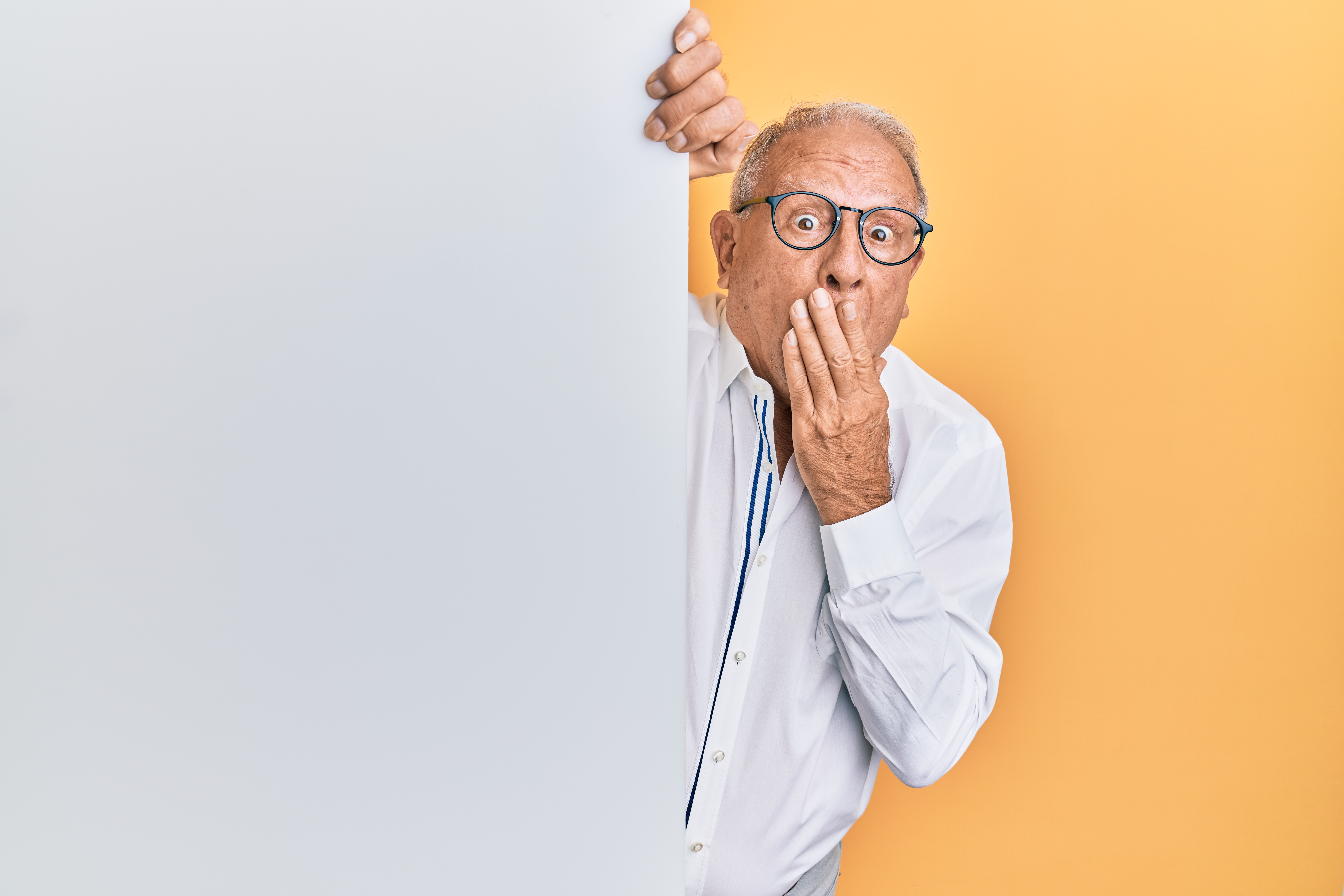 old man in glasses peaking around a white wall  in front of orange background wearing white shirt with hand over mouth looking alarmed