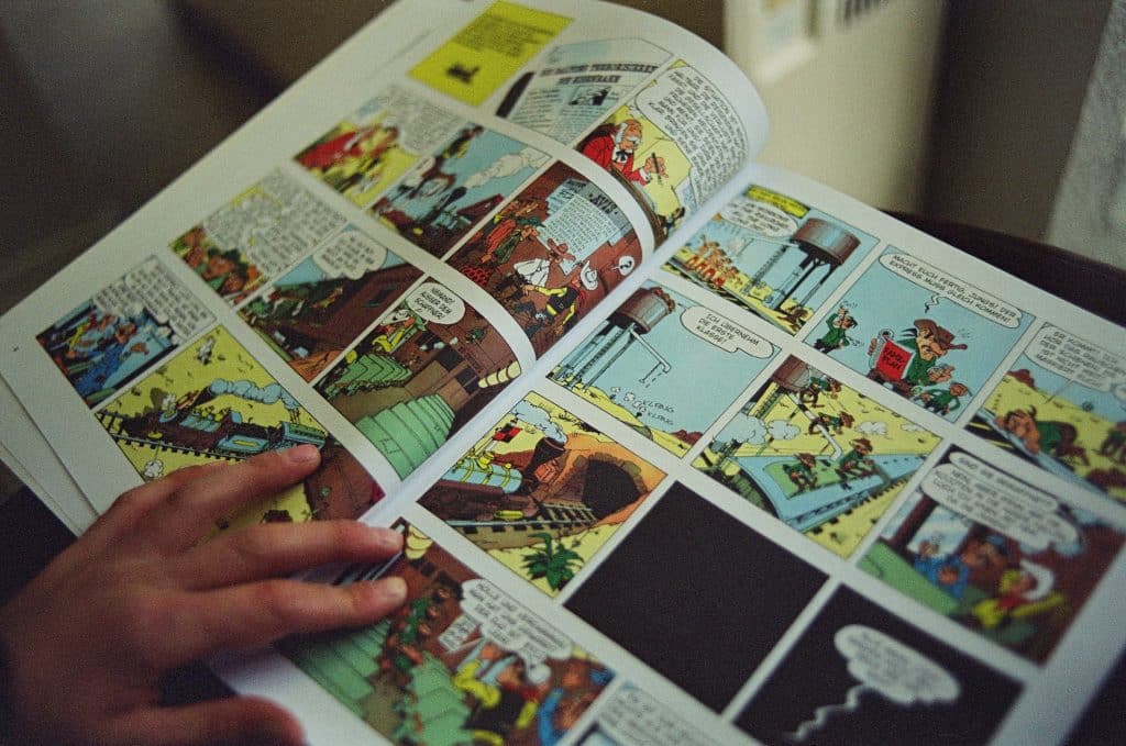 Offset downward view of open comic book with colourful cartoon panes with person holding open the page with left hand