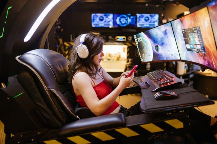 Side profile of Female gamer in red top sitting in black leather chair in front of gaming monitors holding a mobile phone